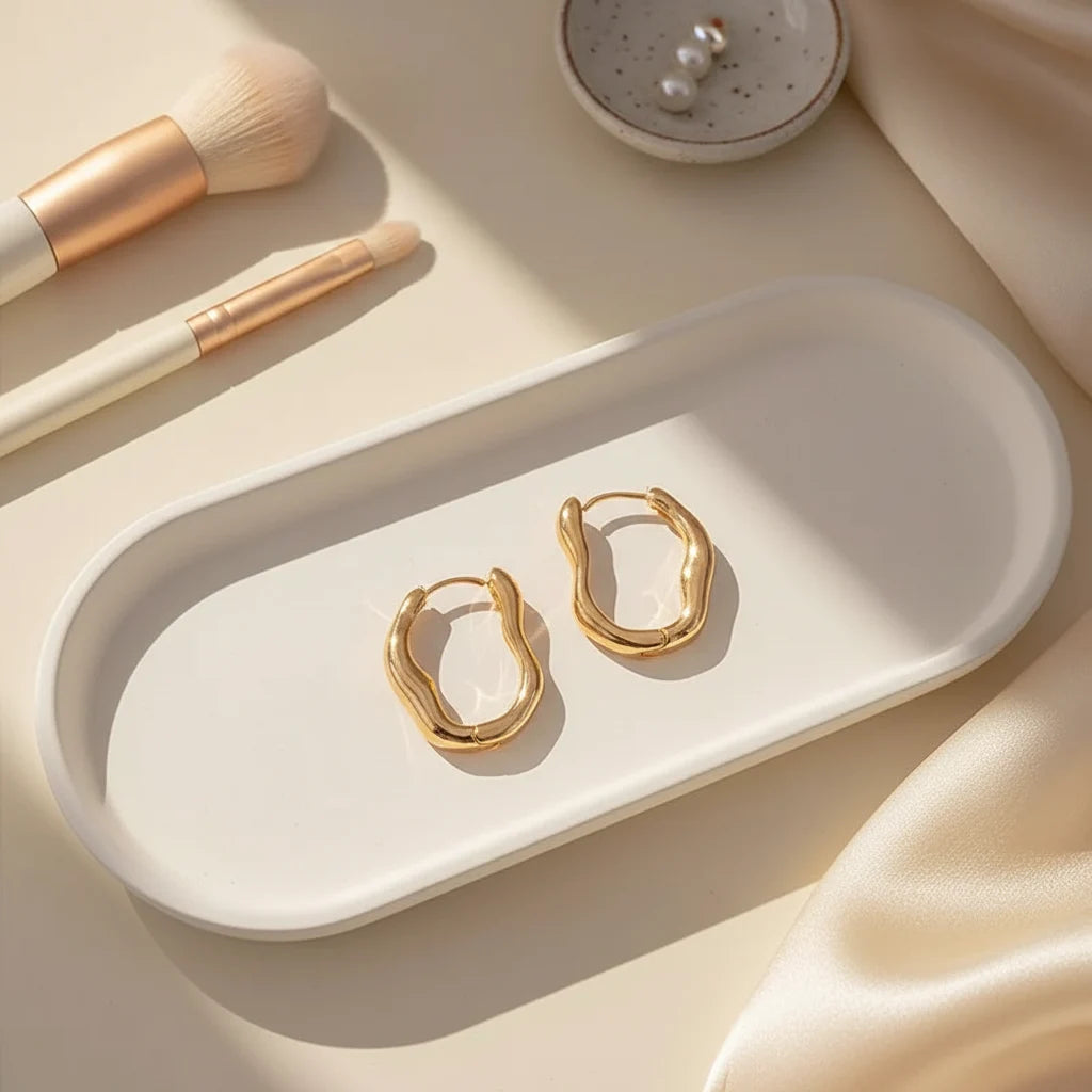 Gold Huggie Hoop Earrings on tray with makeup brushes in soft sunlight | OriaJewel