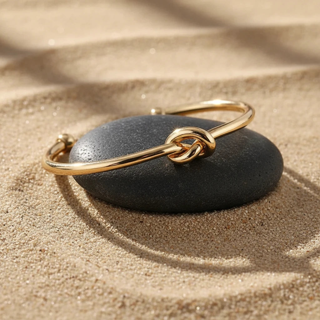 Close-up of a polished Gold Knot Cuff Bracelet resting on a smooth dark stone in warm sand, highlighting the shiny knot detail and open cuff ends | Oria Jewel