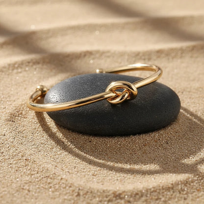 Close-up of a polished Gold Knot Cuff Bracelet resting on a smooth dark stone in warm sand, highlighting the shiny knot detail and open cuff ends | Oria Jewel