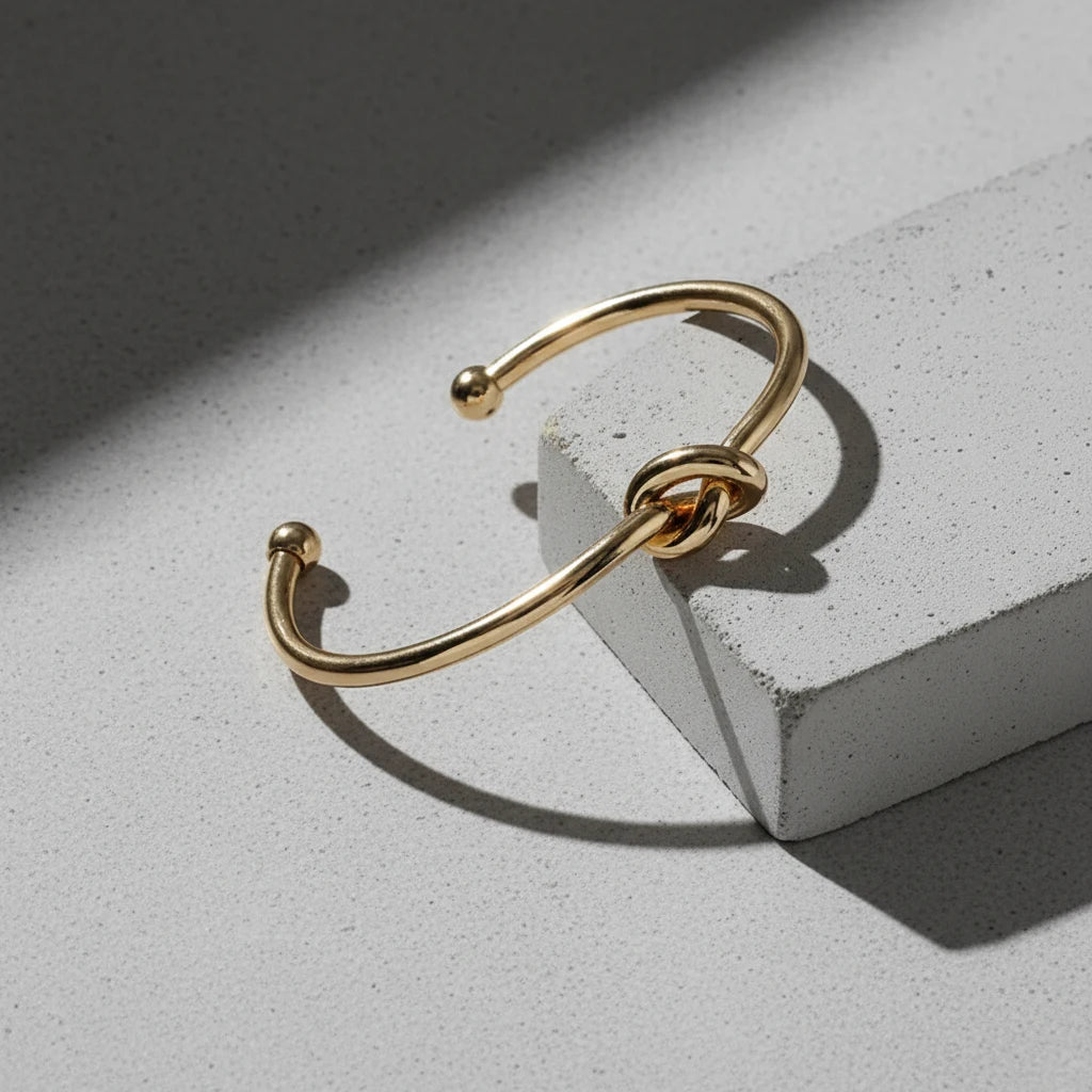 Elegant studio shot of a Gold Knot Cuff Bracelet resting across a light gray concrete block, its polished gold knot detail casting a soft shadow that highlights the minimalist, modern design | Oria Jewel