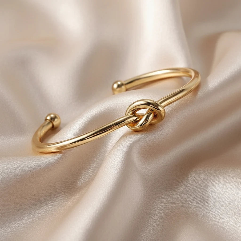 Shiny Gold Knot Cuff Bracelet displayed on soft champagne satin fabric, emphasizing its smooth curved band and delicate central knot design | Oria Jewel