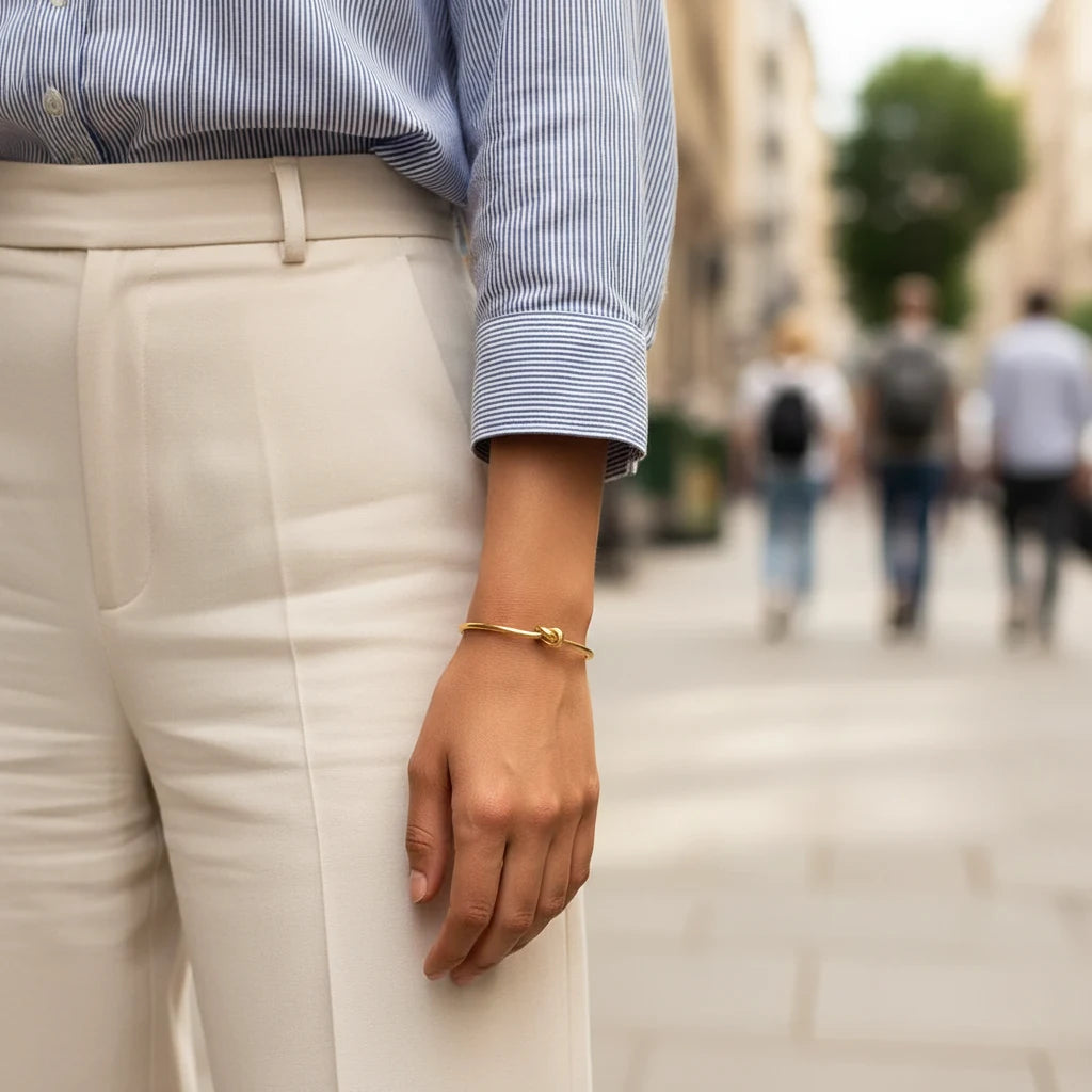 Woman wearing a Gold Knot Cuff Bracelet with white trousers and a striped shirt while walking in the city, bracelet adding a refined touch to her casual outfit | Oria Jewel