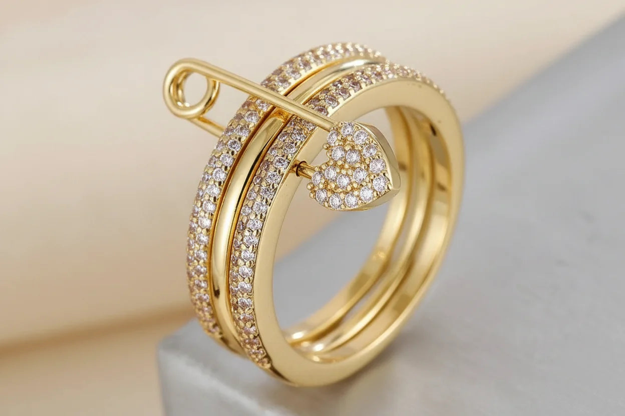 Gold Safety Pin Ring close-up on a neutral surface, gold-tone triple-band look with pavé crystals, safety pin detail across the top and a pavé heart charm, sharp macro focus | OriaJewel
