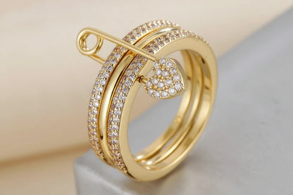 Gold Safety Pin Ring close-up on a neutral surface, gold-tone triple-band look with pavé crystals, safety pin detail across the top and a pavé heart charm, sharp macro focus | OriaJewel