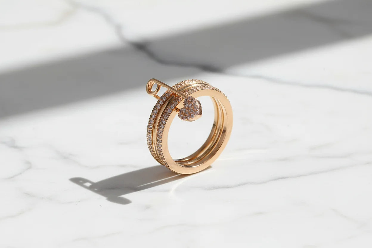 Gold Safety Pin Ring standing on white marble in bright natural light, long clean shadow, gold-tone pavé bands with safety pin motif and sparkling heart charm | OriaJewel