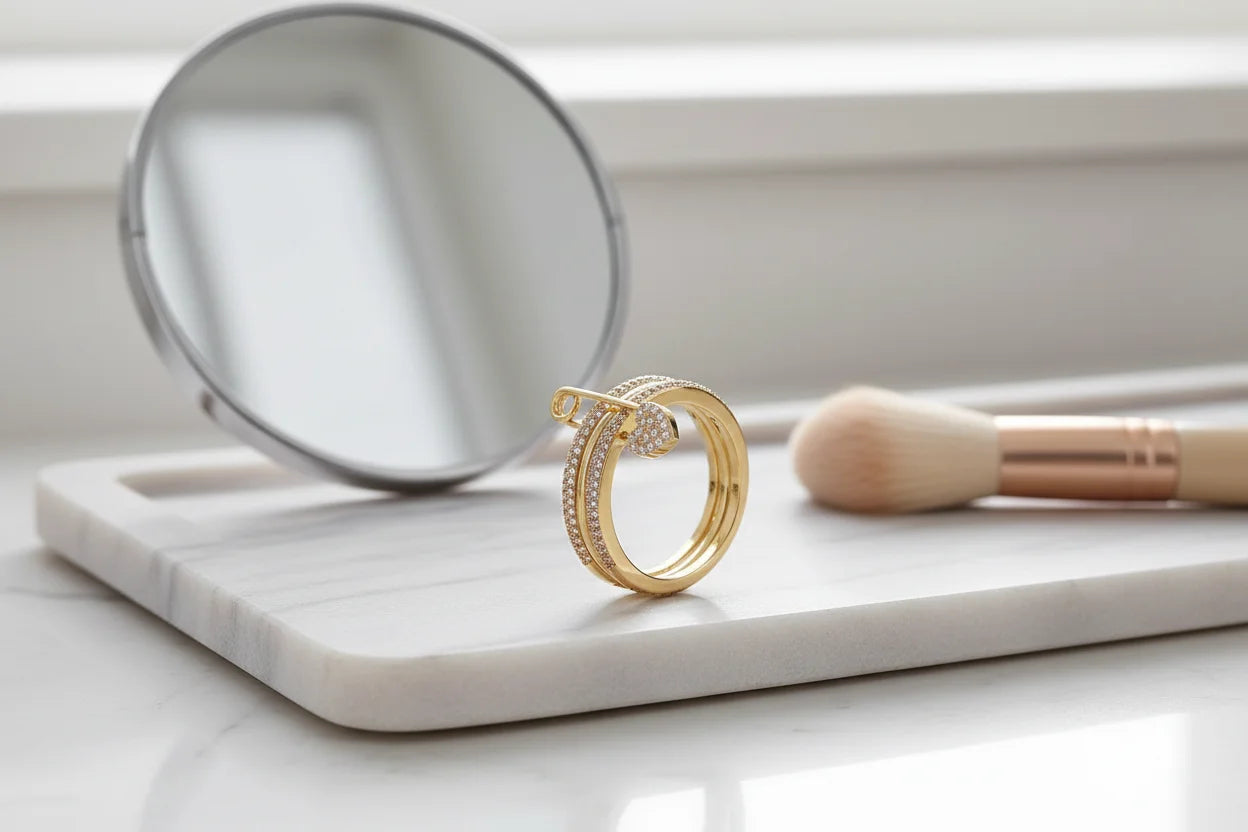 Gold Safety Pin Ring on a marble tray beside a round mirror and makeup brush, clean vanity scene with diffused daylight, focus on safety pin design and pavé heart charm | OriaJewel