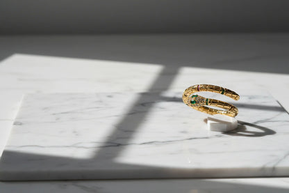 Gold Snake Wrap Ring displayed on white marble under window-grid shadows, side view highlighting the open wrap coil and sparkling snake head with green accents | OriaJewel