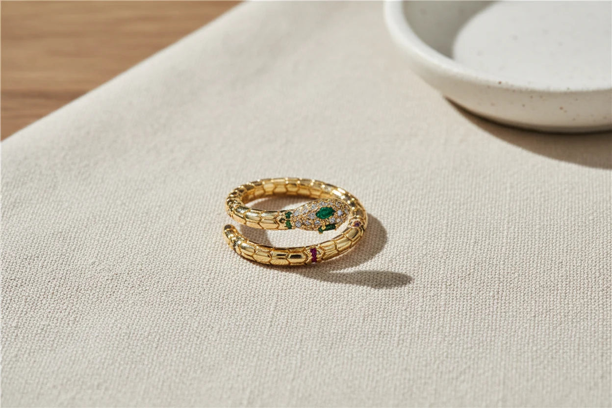 Gold Snake Wrap Ring close-up on textured beige fabric near a ceramic dish, warm natural light emphasizing the gold scale pattern and crystal-studded snake head | OriaJewel