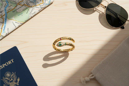 Gold Snake Wrap Ring flat-lay travel scene on a light wood table with a passport, map corner, sunglasses, and a drawstring pouch, featuring a textured snake coil and crystal snake head detail | OriaJewel