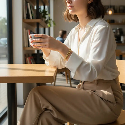 Woman sitting at a café table holding a cup with both hands, wearing a white blouse and beige trousers, with a Gold chain bracelet on her wrist catching the soft window light | Oria Jewel