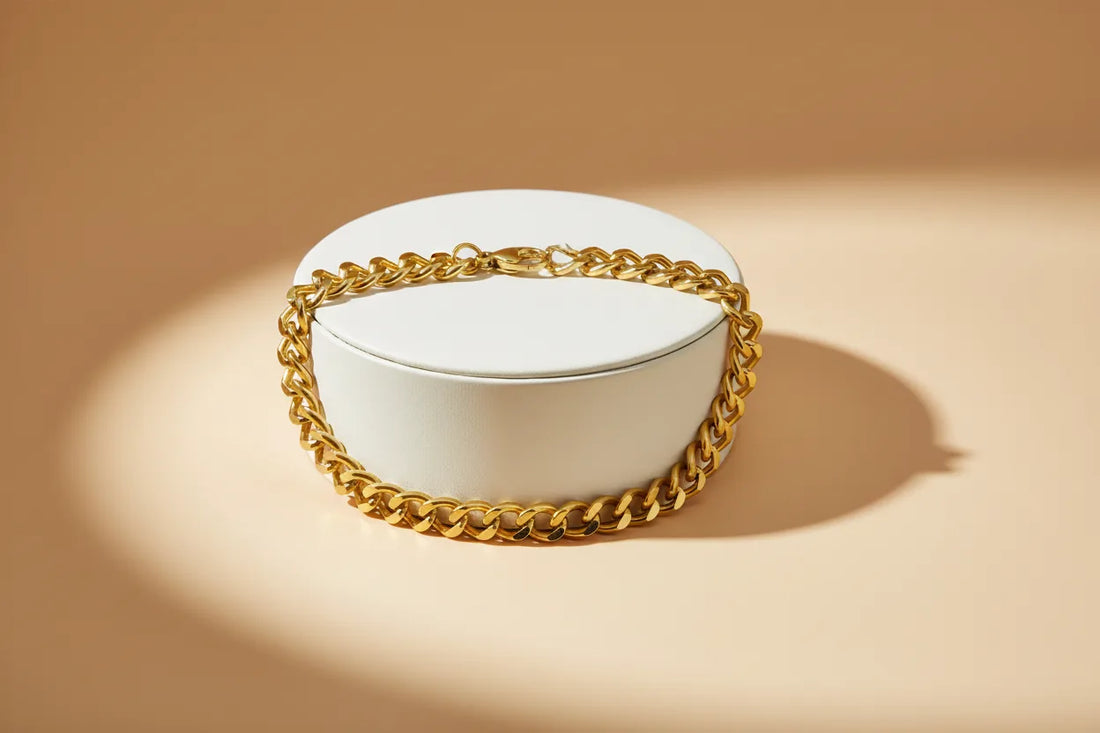 Gold chain bracelet draped around a round white jewelry box on a beige background, highlighted by a soft spotlight to emphasize its polished links and shine | Oria Jewel