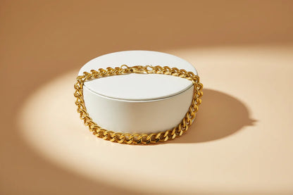 Gold chain bracelet draped around a round white jewelry box on a beige background, highlighted by a soft spotlight to emphasize its polished links and shine | Oria Jewel