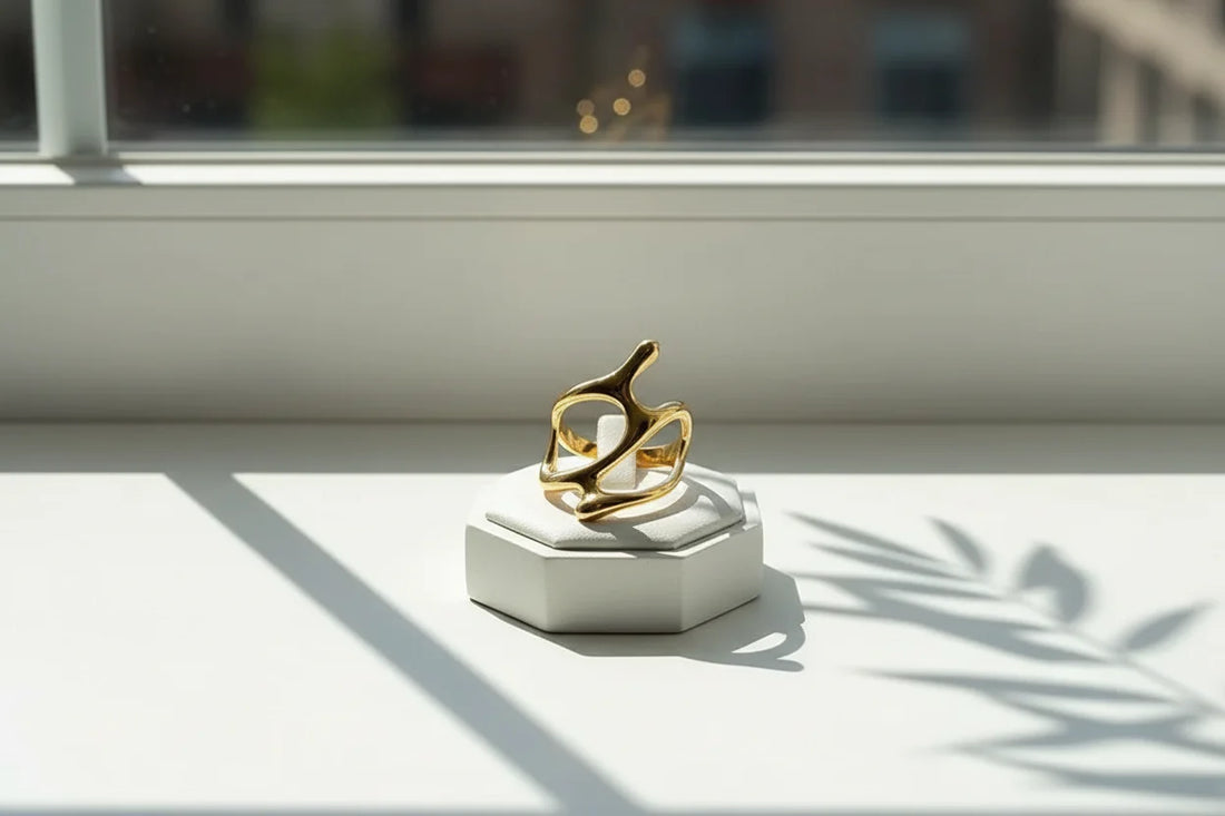 Sunlit product shot of the Gold organic shape ring displayed on a small geometric stand on a windowsill, bright natural light, soft bokeh outside the window, and botanical shadows on the surface | OriaJewel