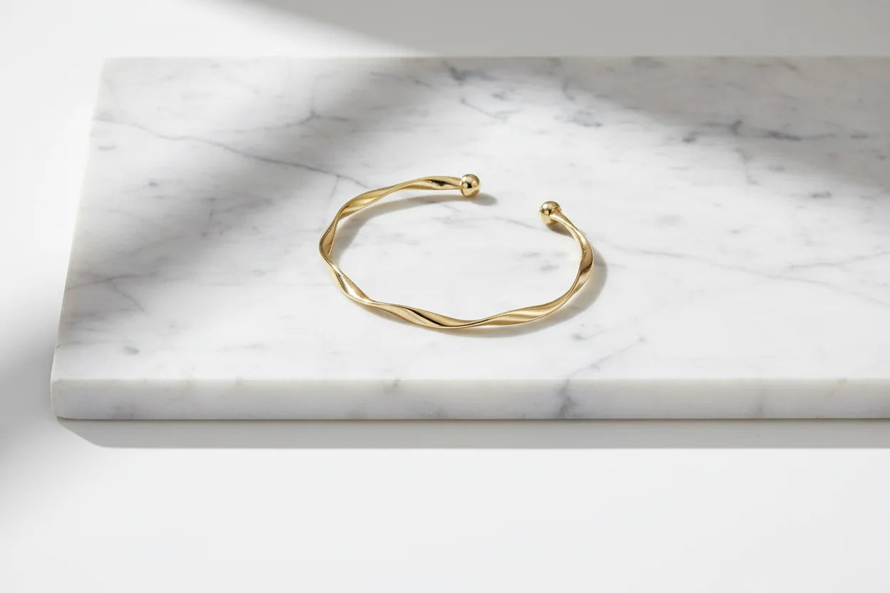 Elegant Gold twisted cuff bracelet placed on a smooth white marble slab, its reflective surface highlighting the spiral design in soft natural light | Oria Jewel