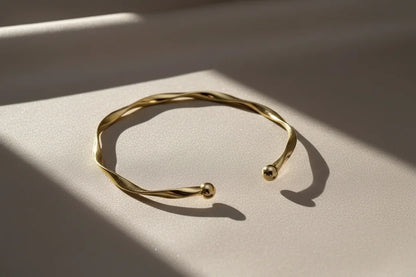 Close-up of Gold twisted cuff bracelet on a neutral fabric background with angled sunlight creating warm shadows that enhance its sculptural twist | Oria Jewel
