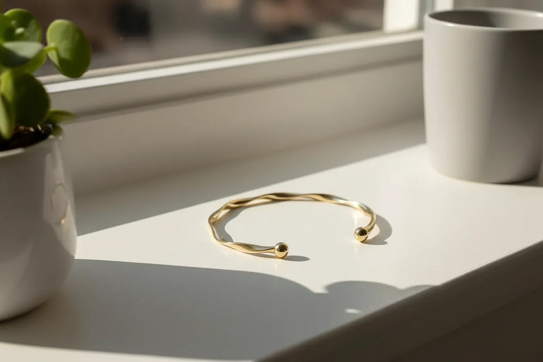 Gold twisted cuff bracelet displayed on a bright window ledge beside a ceramic pot, with sunlight streaming in to emphasize its smooth metallic curves | Oria Jewel