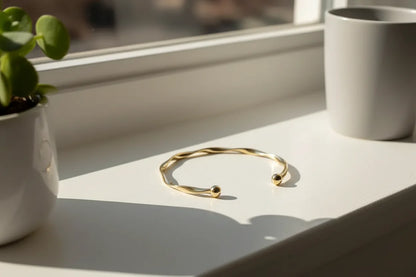 Gold twisted cuff bracelet displayed on a bright window ledge beside a ceramic pot, with sunlight streaming in to emphasize its smooth metallic curves | Oria Jewel