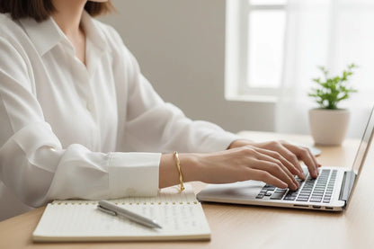 Woman typing on a laptop at a minimal workspace, wearing a Gold twisted cuff bracelet that adds a polished accent to her white blouse and modern desk setup | Oria Jewel
