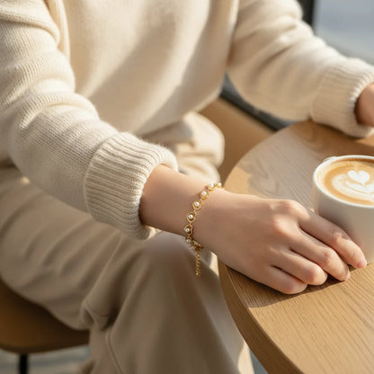 Woman in a cozy cream sweater holding a latte at a café table, with the Golden Pearl Heart Chain Bracelet sparkling on her wrist, its tiny pearl-filled hearts catching the warm morning light | Oria Jewel