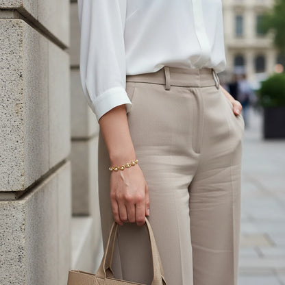 Chic street-style look with a woman in a white blouse and tailored beige trousers holding a structured tote bag, the Golden Pearl Heart Chain Bracelet adding a delicate gold and pearl accent to her wrist | Oria Jewel