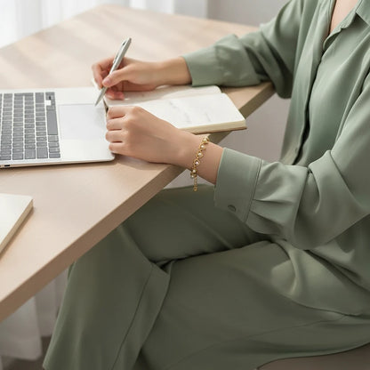 Minimal home office scene with a woman in a soft sage-green set writing in a notebook beside her laptop, the Golden Pearl Heart Chain Bracelet wrapping her wrist in dainty heart links and pearls | Oria Jewel