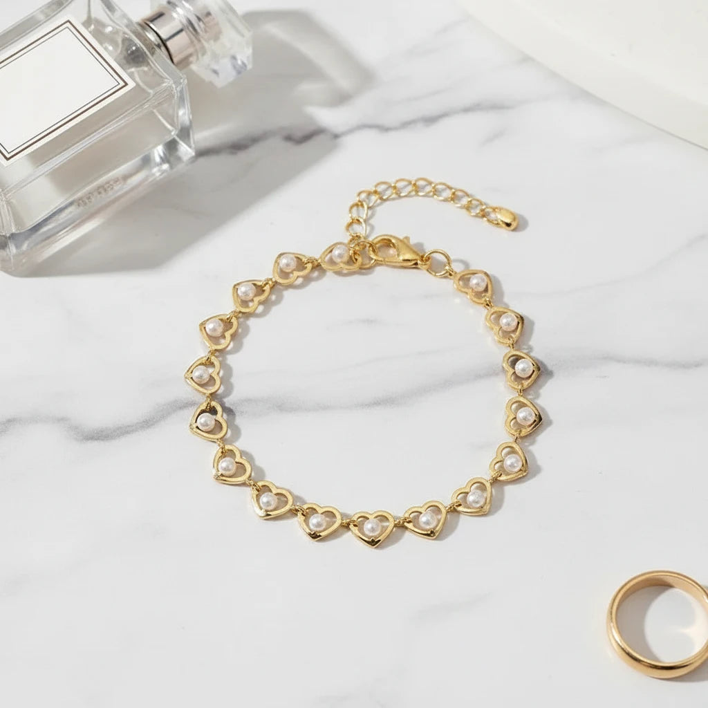 Close-up flat lay of the Golden Pearl Heart Chain Bracelet arranged in a circle on white marble beside a clear perfume bottle and gold ring, highlighting its dainty pearl-filled hearts and adjustable clasp | Oria Jewel