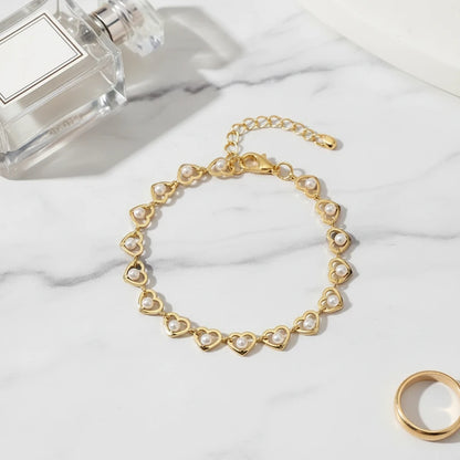 Close-up flat lay of the Golden Pearl Heart Chain Bracelet arranged in a circle on white marble beside a clear perfume bottle and gold ring, highlighting its dainty pearl-filled hearts and adjustable clasp | Oria Jewel