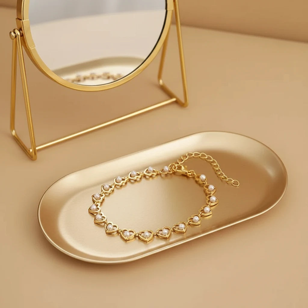 Golden Pearl Heart Chain Bracelet resting on an oval gold tray in front of a round vanity mirror, the reflective surface enhancing the shine of the polished hearts and tiny pearls | Oria Jewel