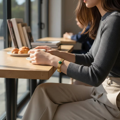 Woman sitting at a café table in soft daylight, holding a cup with both hands while a Green Gemstone Cuff Bracelet in gold-tone metal and an oval green stone is clearly visible on her wrist, styled with a ribbed dark grey top and beige trousers for a relaxed, modern look | Oria Jewel