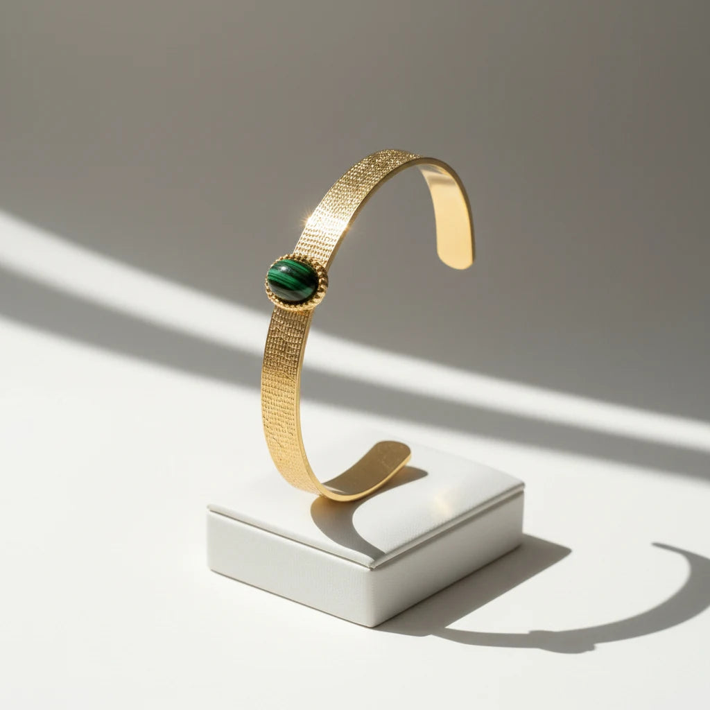 Minimalist hero shot of the Green Gemstone Cuff Bracelet standing upright on a small white plinth, with bright sunlight and soft shadows emphasizing the textured gold band and striped green stone centerpiece | Oria Jewel
