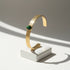 Minimalist hero shot of the Green Gemstone Cuff Bracelet standing upright on a small white plinth, with bright sunlight and soft shadows emphasizing the textured gold band and striped green stone centerpiece | Oria Jewel