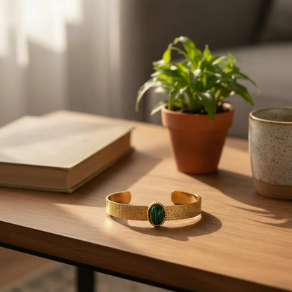 Lifestyle image of the Green Gemstone Cuff Bracelet placed on a warm wooden coffee table next to a potted plant, book, and speckled mug, suggesting a cozy, slow-morning at-home vibe | Oria Jewel
