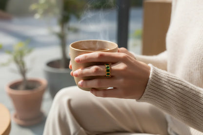 Green stone band ring on a hand holding a warm ceramic cup, soft morning daylight and cozy neutral outfit, focus on the gold-tone band and green stones | OriaJewel