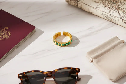 Green stone band ring flat lay on a bright marble surface with travel accessories, sunglasses and a pouch nearby, sharp detail on the gold-tone band and green stones | OriaJewel