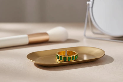 Green stone band ring placed on a brushed gold tray near beauty tools and a mirror, warm editorial lighting and close focus on the gold-tone finish and green stones | OriaJewel