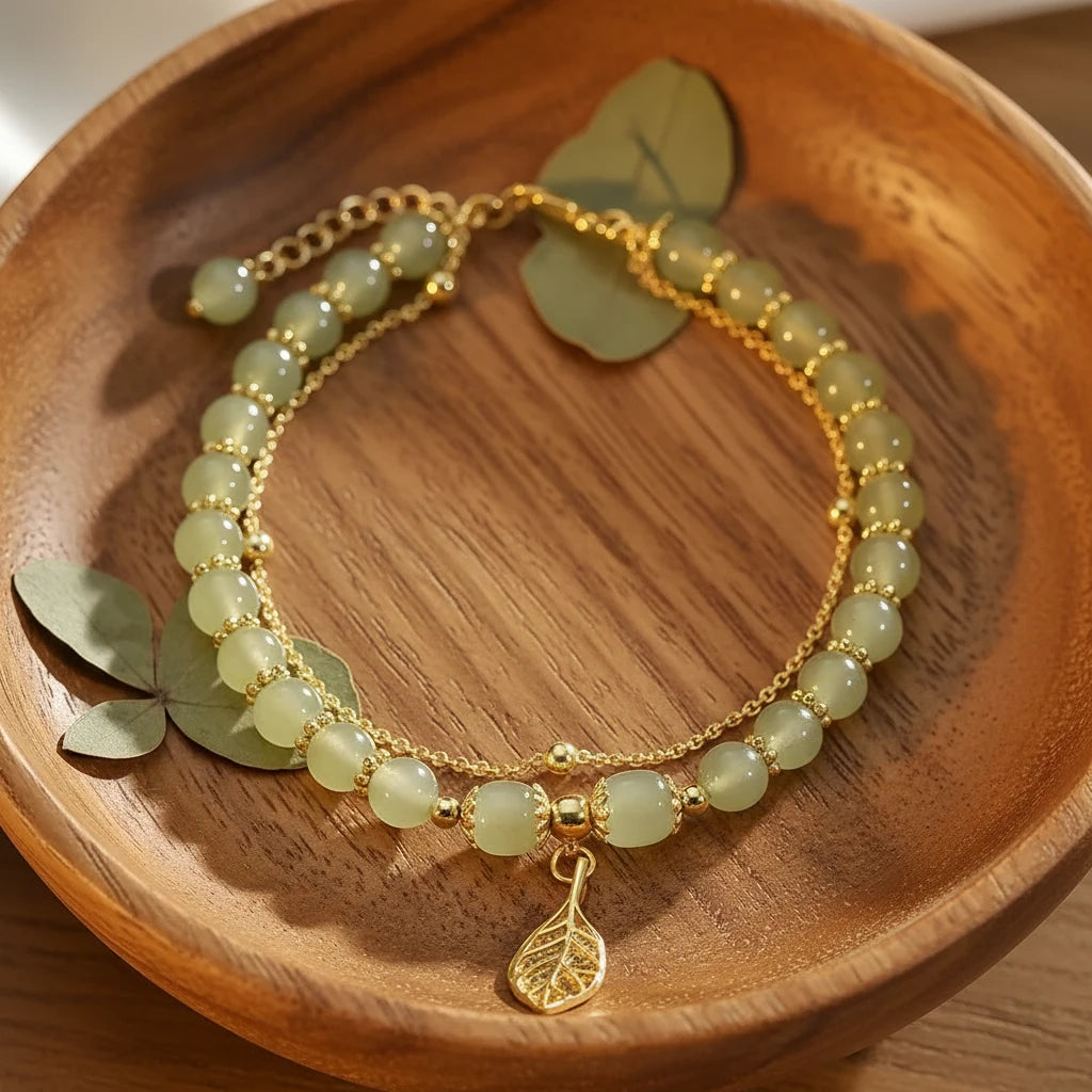 Jade Leaf adjustable charm bracelet displayed in a wooden dish with soft sunlight highlighting the gold chain and leaf charm | Oria Jewel