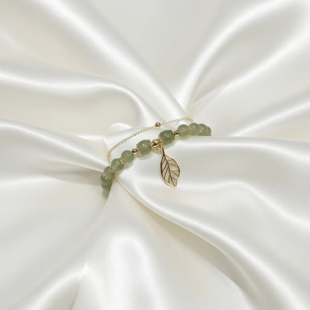 Jade Leaf adjustable charm bracelet arranged on white satin fabric, gold leaf charm and green beads catching the light elegantly | Oria Jewel
