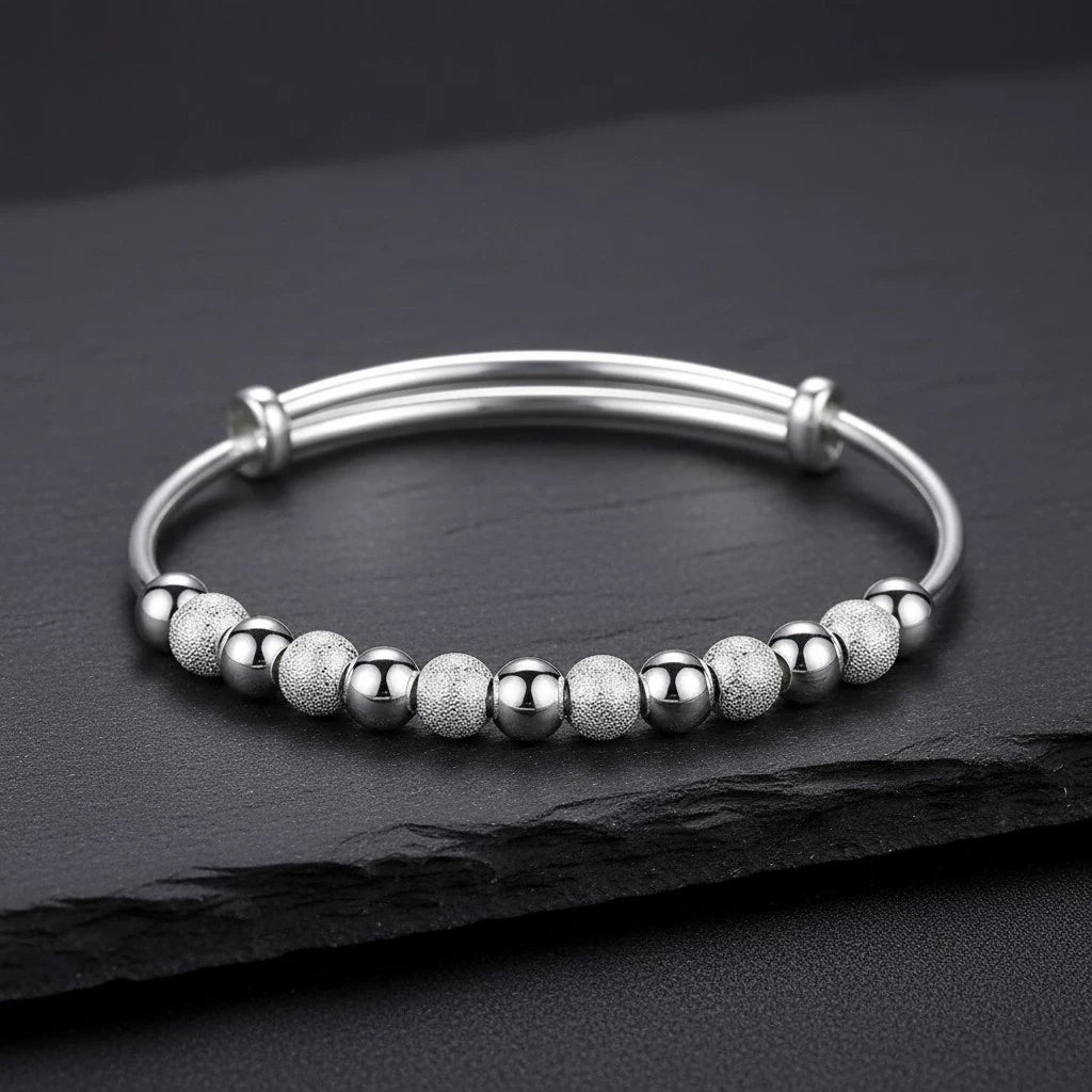 The Marvella Beaded Charm Cuff laid flat on a textured slate surface, showcasing the row of alternating smooth and sandblasted beads along a polished open cuff for an elegant, versatile style | Oria Jewel

