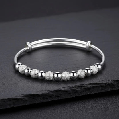 The Marvella Beaded Charm Cuff laid flat on a textured slate surface, showcasing the row of alternating smooth and sandblasted beads along a polished open cuff for an elegant, versatile style | Oria Jewel

