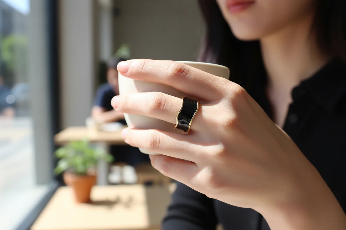 Midnight Black Open Wave Ring worn on a hand holding a white coffee cup in a bright café, glossy black wave band with gold-tone edges in soft daylight | OriaJewel