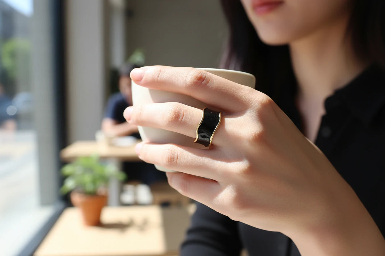 Midnight Black Open Wave Ring worn on a hand holding a white coffee cup in a bright café, glossy black wave band with gold-tone edges in soft daylight | OriaJewel