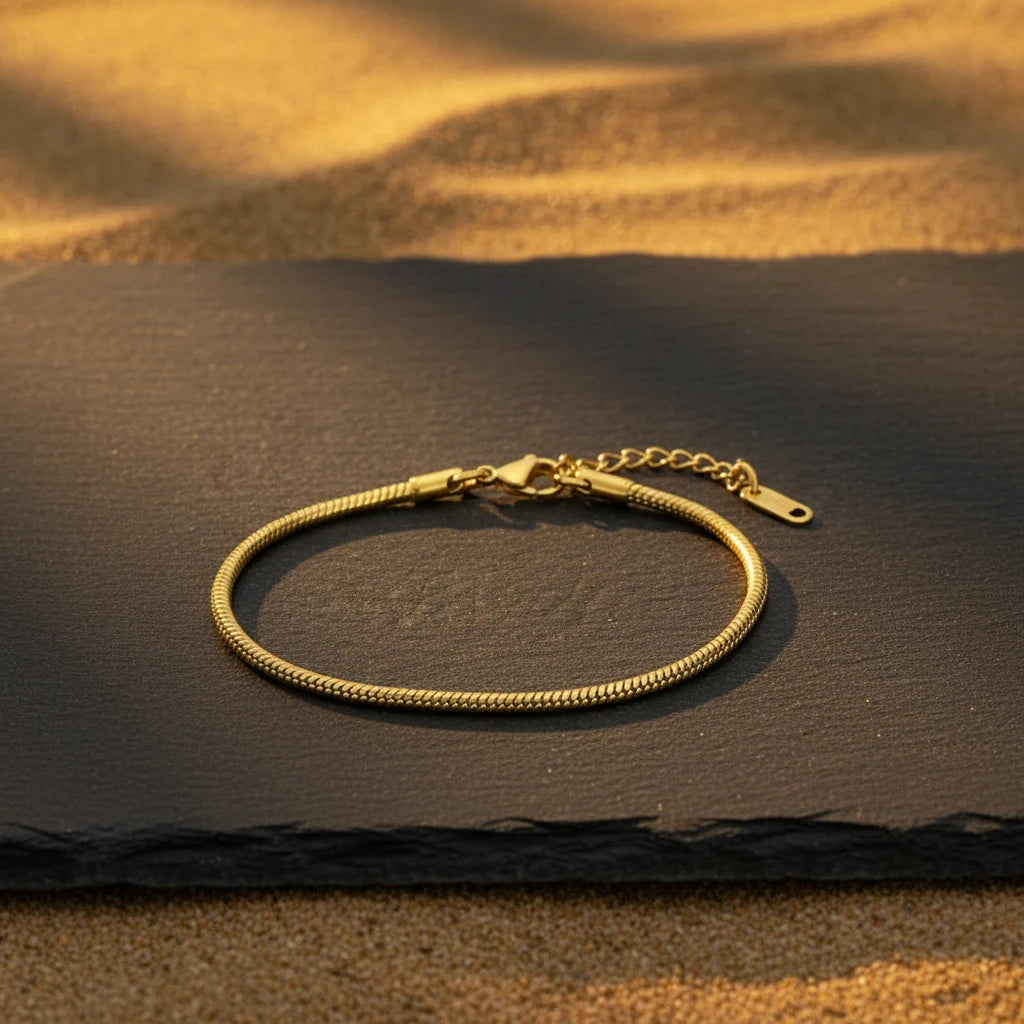Minimalist Gold Chain Bracelet lying on a dark slate tray, surrounded by softly lit golden sand texture | Oria Jewel