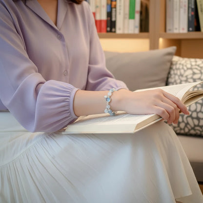 Woman reading on a sofa wearing a pastel blouse and white skirt, showing the delicate Moonstone Glow Charm Bracelet on her wrist | Oria Jewel