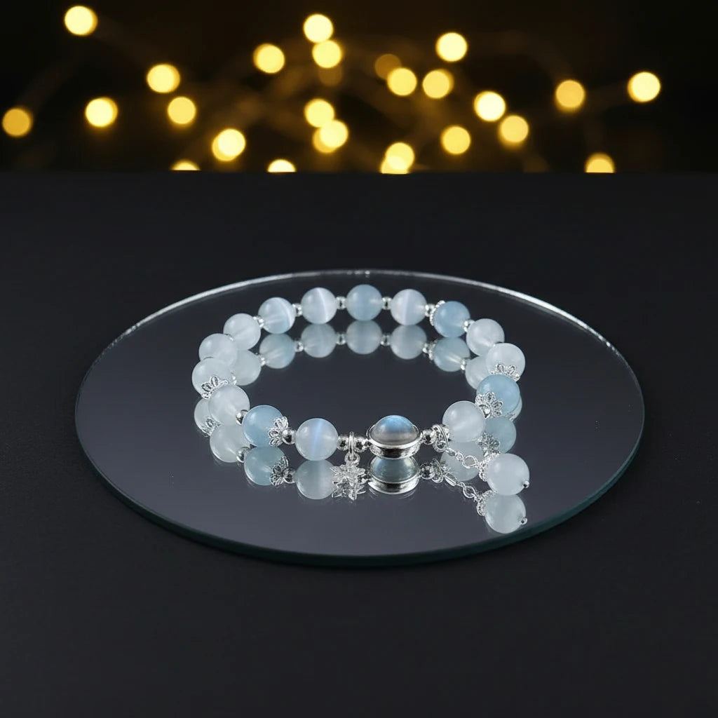 Moonstone Glow Charm Bracelet resting on a round white display with flowing fabric, featuring a glowing moonstone, butterfly charm, and dangling pearl | Oria Jewel