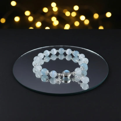 Moonstone Glow Charm Bracelet resting on a round white display with flowing fabric, featuring a glowing moonstone, butterfly charm, and dangling pearl | Oria Jewel