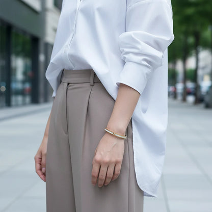 Woman walking in the city wearing a crisp white shirt and taupe tailored trousers, with the Mother of Pearl Gold Bangle shining on her wrist, capturing a chic everyday street-style look | Oria Jewel