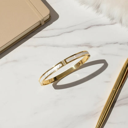 Minimal flat-lay of the Mother of Pearl Gold Bangle on a white marble surface, showing the smooth gold band and glossy mother-of-pearl inlay beside a beige notebook, gold pen, and fluffy detail for an elegant office vibe | Oria Jewel