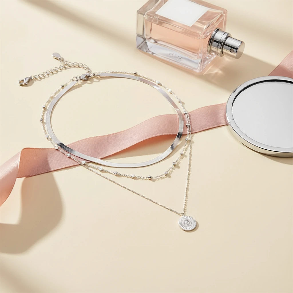 Multi Layer Silver Circle Chain Necklace arranged beside perfume bottle and pink ribbon on beige background | Oria Jewel