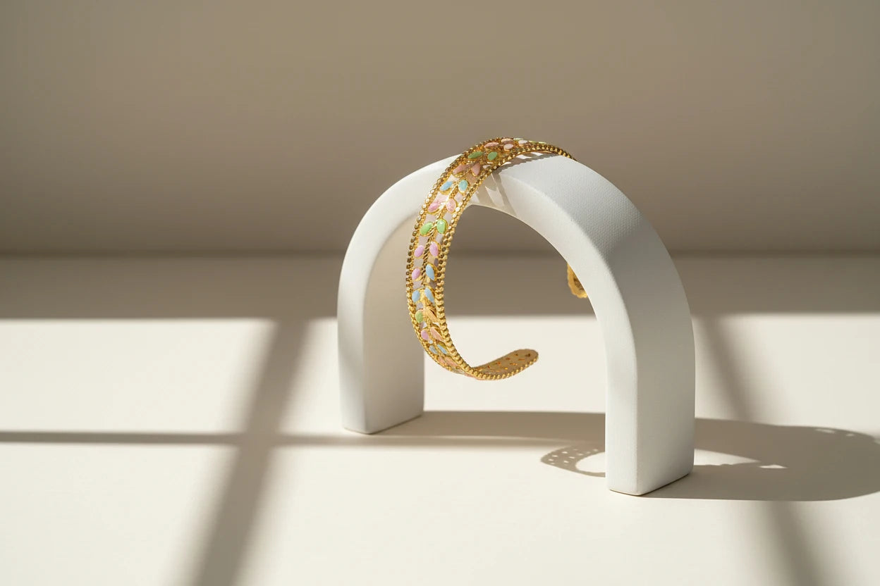 Minimal still life of the Pastel Leaf Garden Cuff Bracelet draped over a white arched stand, sunlight casting soft window-frame shadows to highlight the pastel leaf details and textured gold edges | Oria Jewel