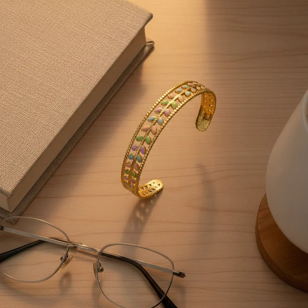 Cozy desk scene with the Pastel Leaf Garden Cuff Bracelet balanced on its edge beside a book, glasses and a mug on a wooden surface, highlighting the intricate pastel leaf design and scalloped gold border | Oria Jewel